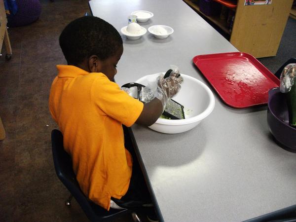 A young child wearing plastic gloves uses a grater to grate zucchini in a large bowl.